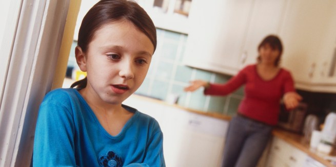 Girl standing in doorway of kitchen, arms folded, woman standing behind, arms extended.